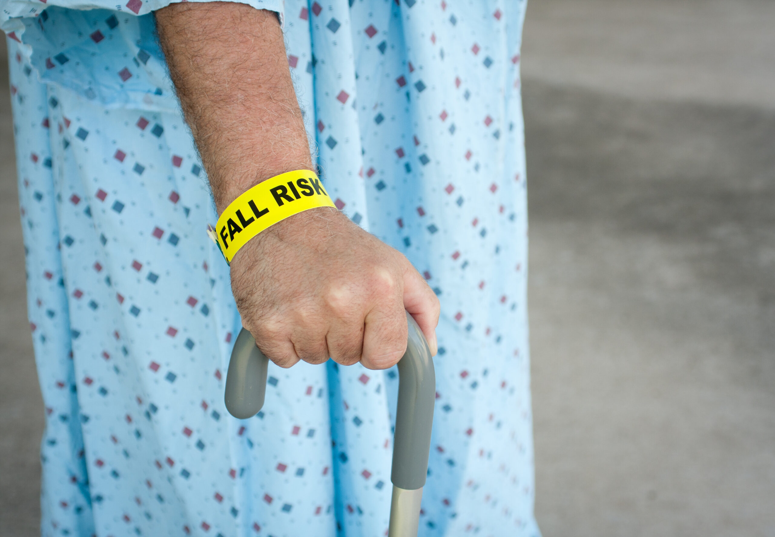 An elderly man wearing a fall risk bracelet around his wrist at the hospital. Wearing a blue gown and walking with a cane.