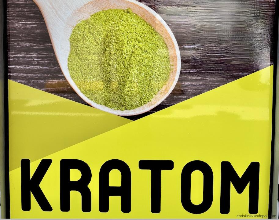 Sign spelling the word kratom in black letters on neon yellow background with a spoon holding green powdered material